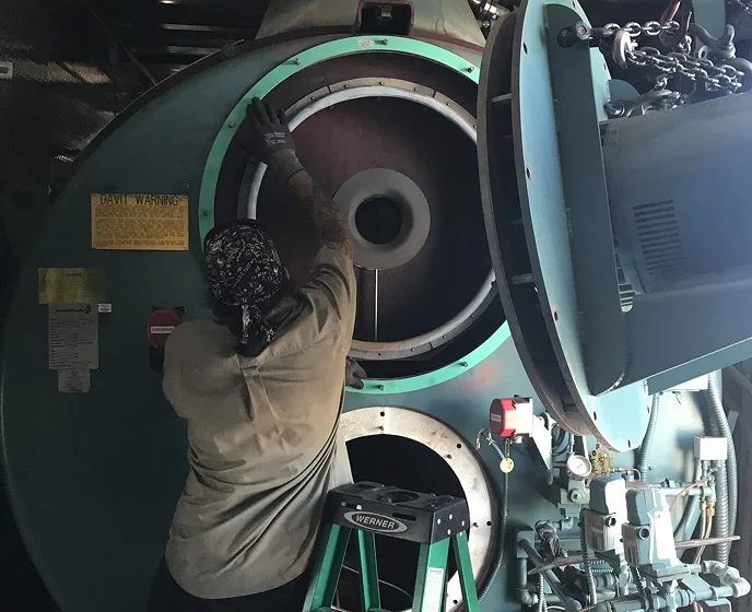 Technician working on a large, heavy-duty industrial fan or blower unit, standing on a ladder.