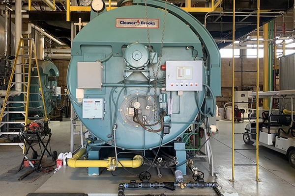 A wide shot of a large, light blue Cleaver-Brooks industrial boiler in a mechanical room, surrounded by pipes, gauges, and other equipment.